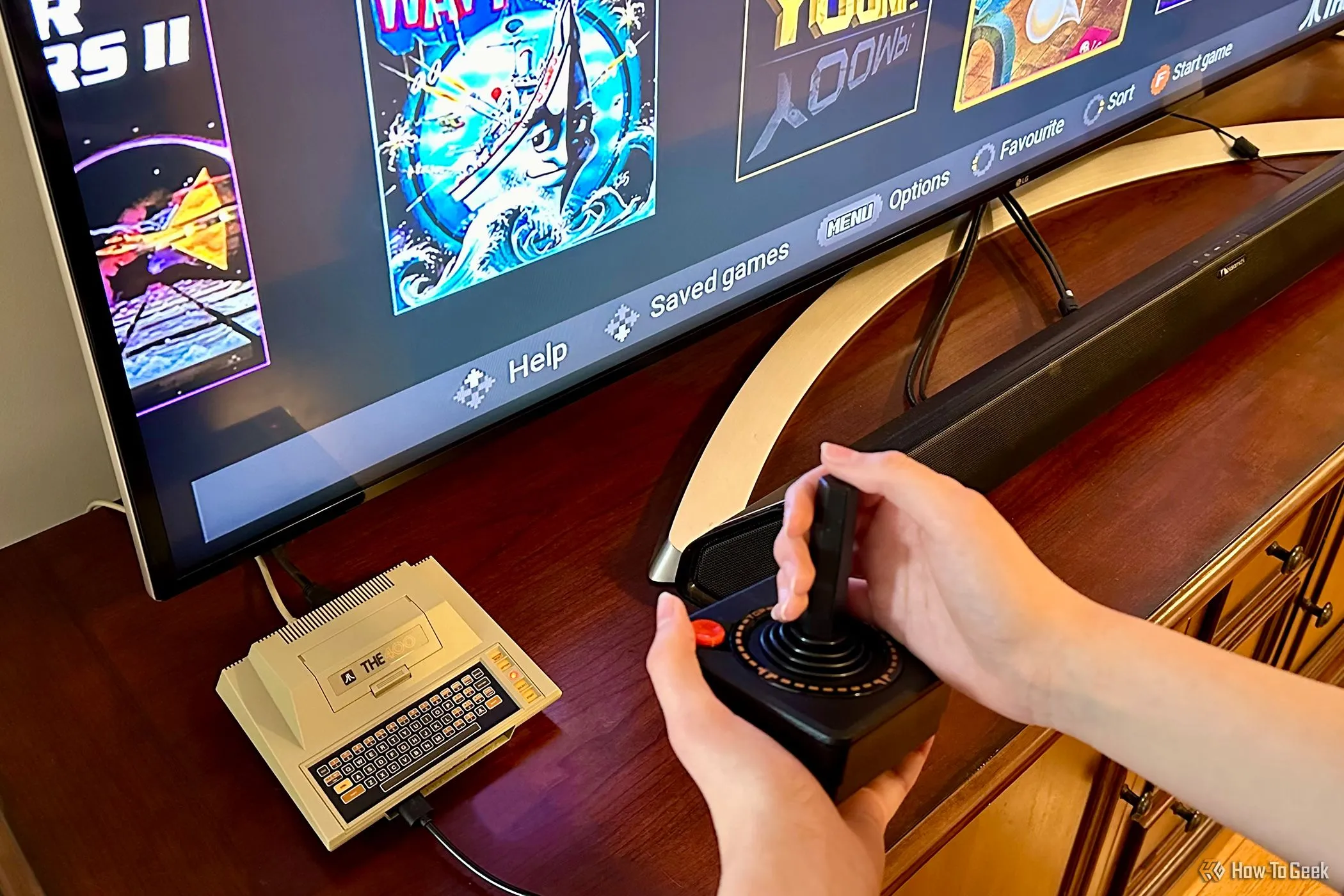 Bộ giả lập THE400 Mini bên cạnh TV, với bàn tay đang cầm cần điều khiển joystick, thiết bị chơi game.