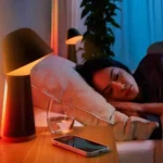 Một người phụ nữ đang ngủ say trên giường cạnh đèn thông minh và điện thoại thông minh, minh họa việc sử dụng điện thoại để hỗ trợ giấc ngủ chất lượng.