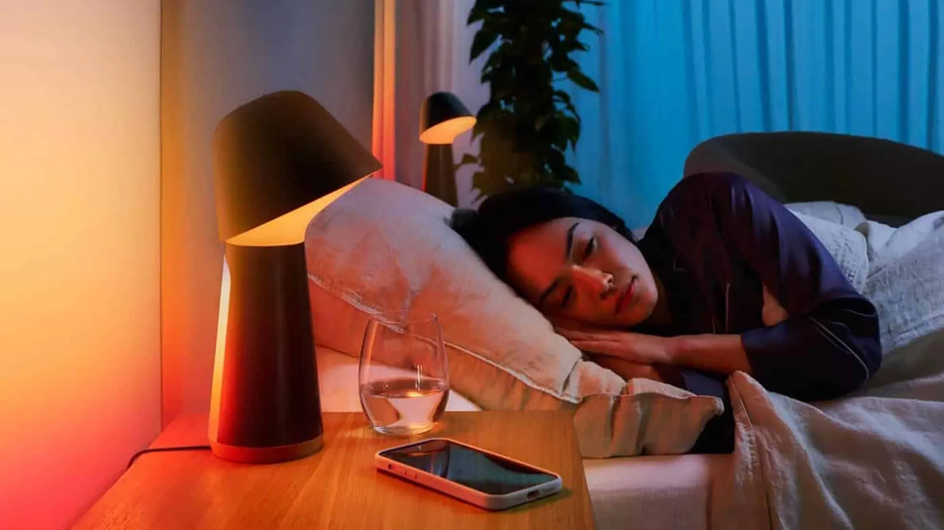 Một người phụ nữ đang ngủ say trên giường cạnh đèn thông minh và điện thoại thông minh, minh họa việc sử dụng điện thoại để hỗ trợ giấc ngủ chất lượng.