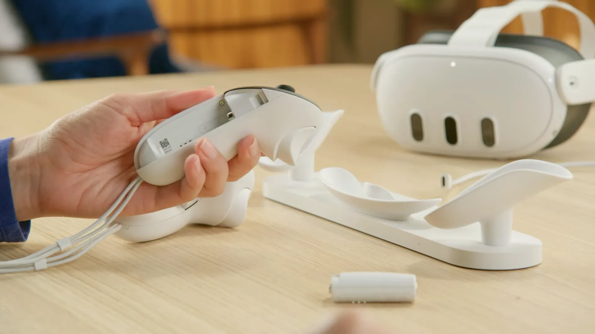 Một tay đang cầm bộ điều khiển Meta Quest 3 Touch Plus với pin sạc, và tai nghe Meta Quest 3 cùng dock sạc ở phía sau.