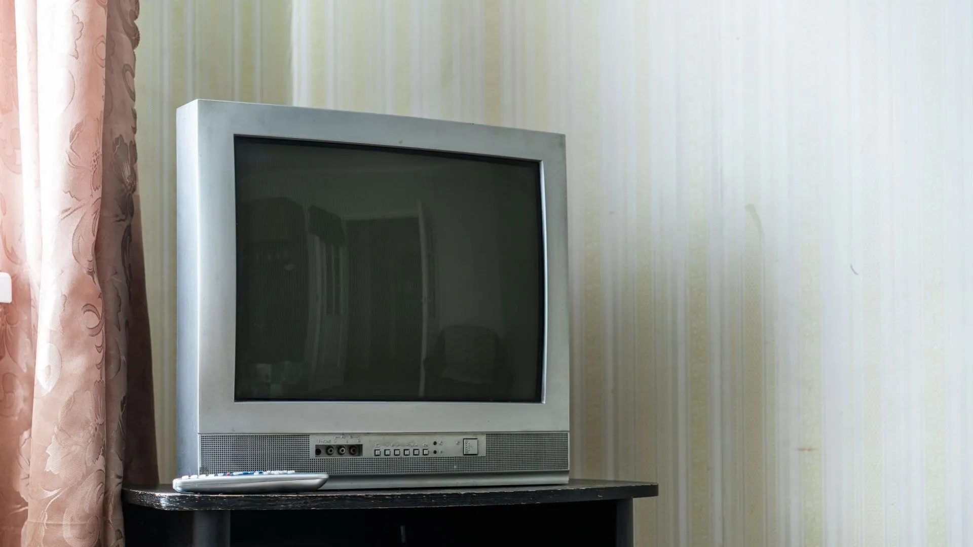 TV CRT kiểu cũ đặt trên bàn tối, trong phòng có rèm cửa và hình nền họa tiết. Điều khiển từ xa đặt cạnh TV, tạo cảm giác hoài niệm.