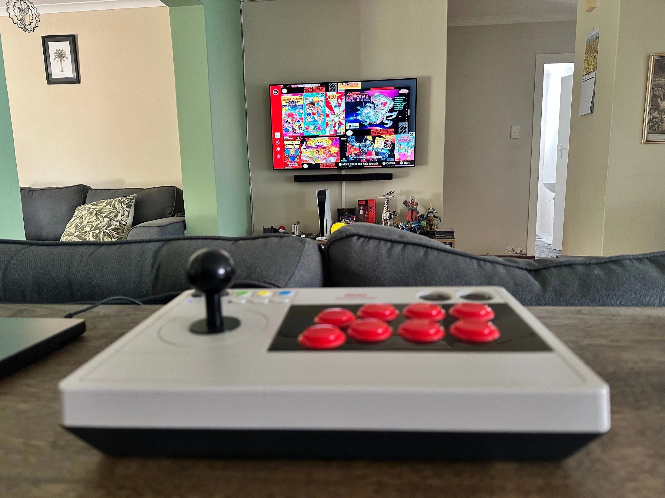 Cần điều khiển arcade 8bitDo màu xám, đặt trước màn hình hiển thị các game Nintendo SNES cổ điển.