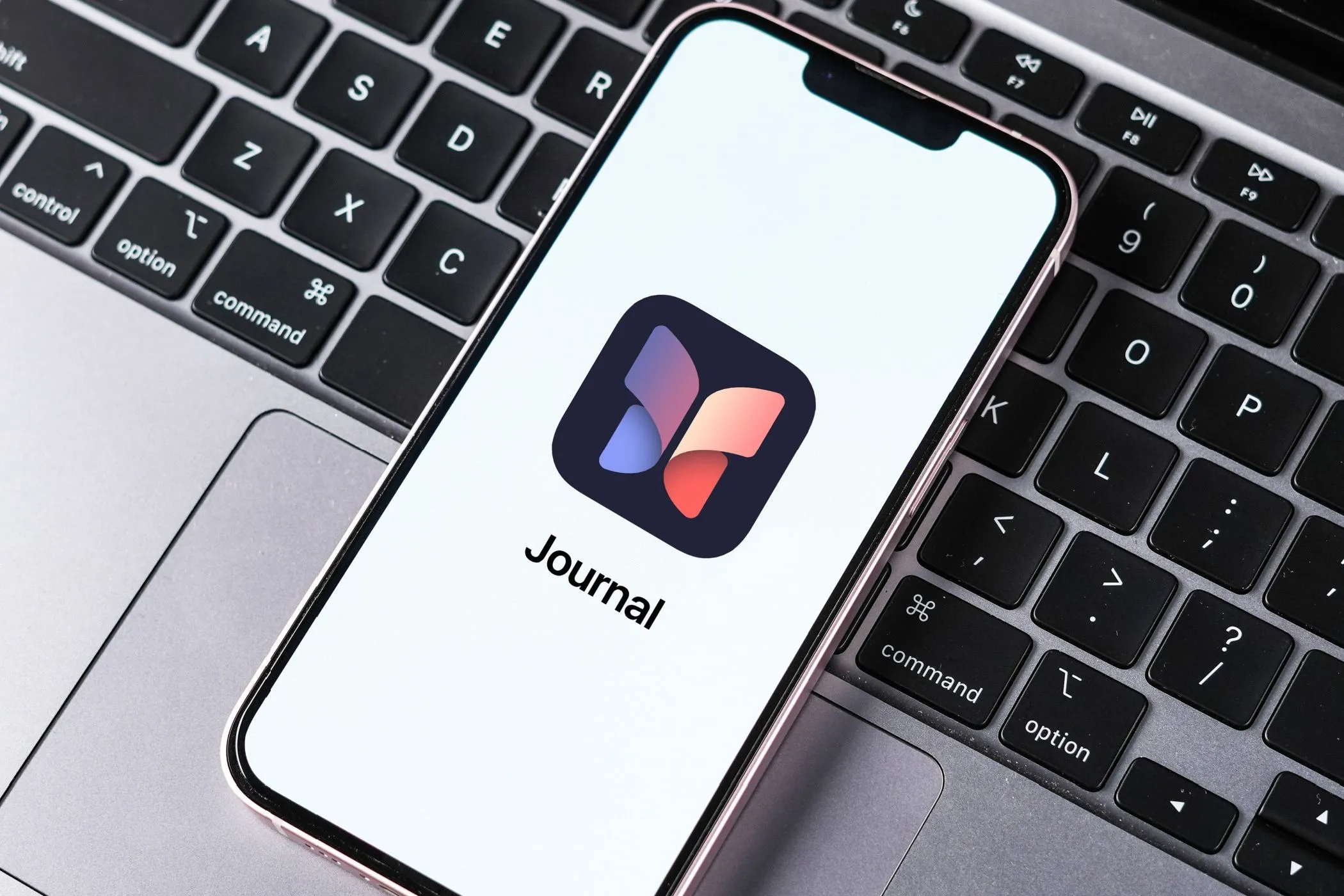 Màn hình iPhone hiển thị ứng dụng Journal của Apple đặt trên bàn phím MacBook, so sánh với nhật ký truyền thống