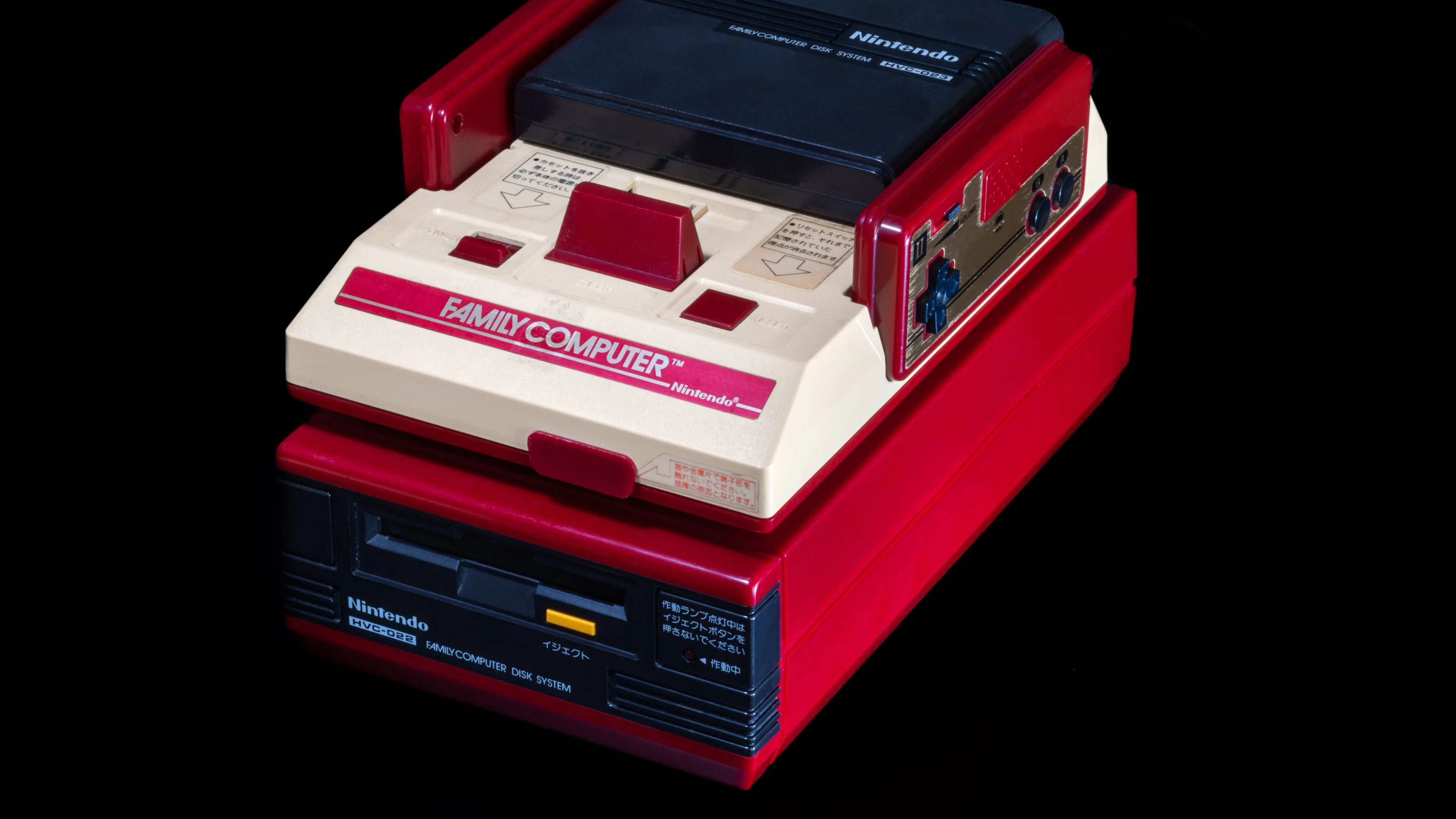 Máy Nintendo Famicom màu đỏ trắng kết nối với hệ thống Famicom Disk System màu vàng, hiển thị khả năng mở rộng của Famicom.